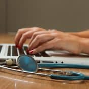prim-plan al unei persoane care tastează pe tastatura unui laptop, cu un stetoscop lângă el